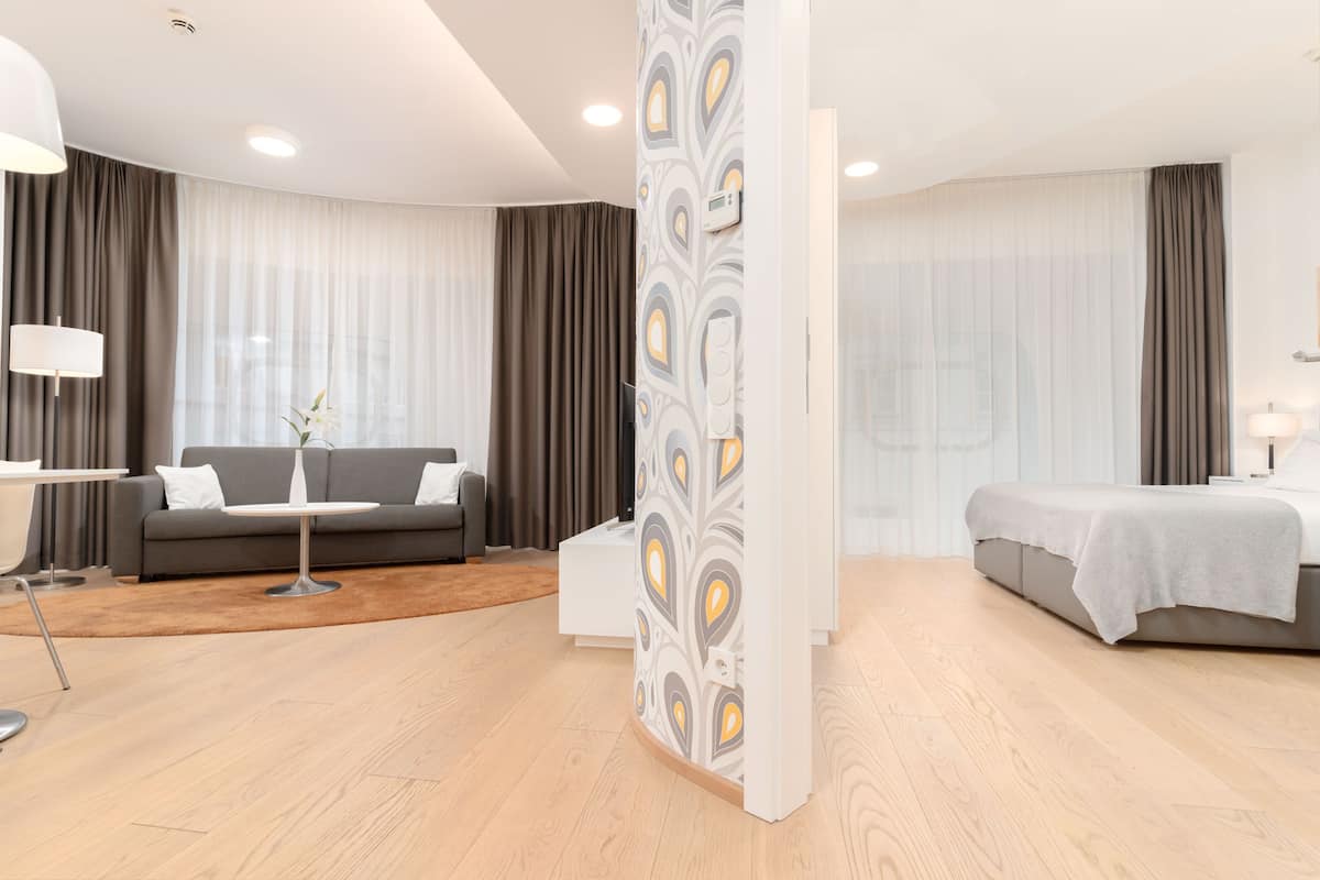 Quarto de hotel moderno com um sofá cinzento, uma mesa redonda pequena, uma cama com cabeceira cinzenta e um divisório branco com padrão decorativo.