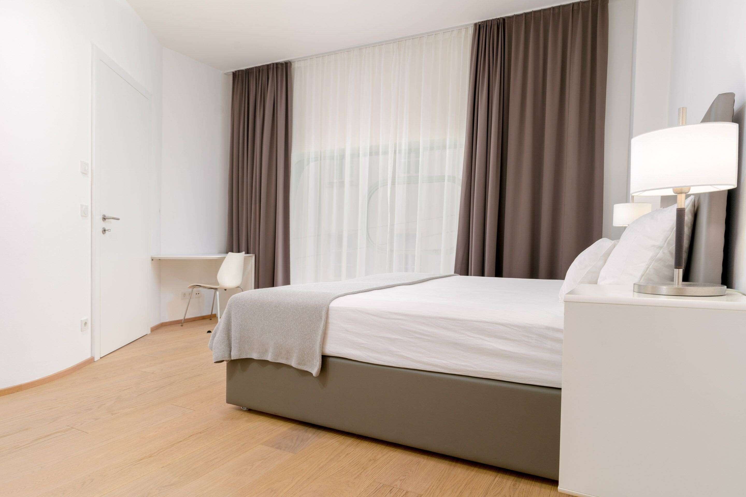 A modern hotel room with a bed, bedside table, lamp, and a desk.