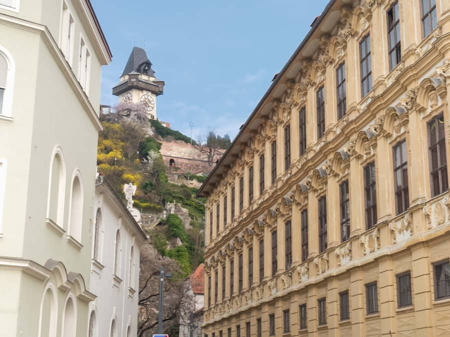 Ein historisches Gebäude mit klassischer Architektur, einem Glockenturm und Blick auf einen bewachsenen Hügel.