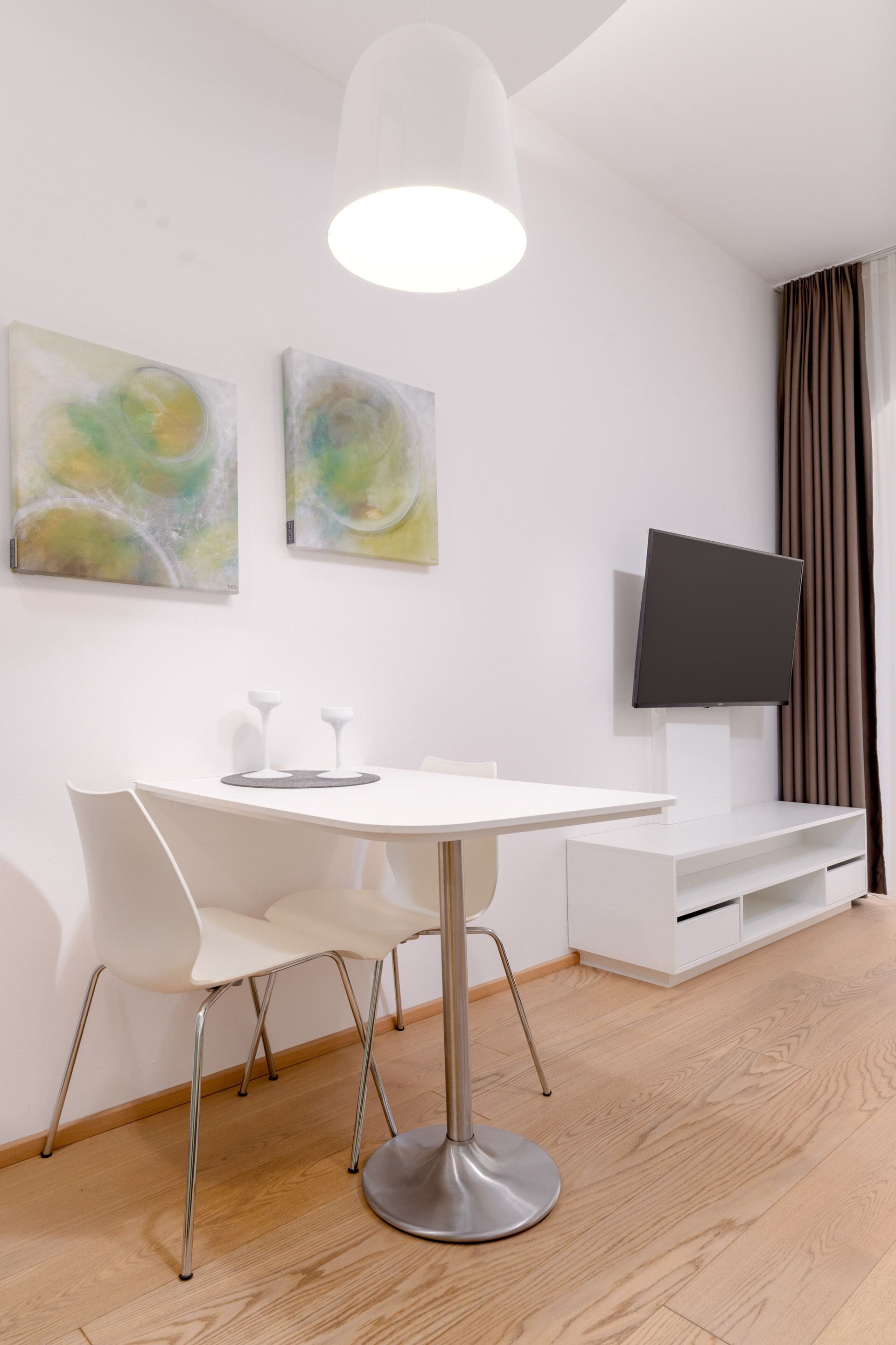 Una habitación de hotel moderna con una mesa de comedor redonda, sillas blancas, un televisor de pantalla plana y pinturas abstractas en la pared.