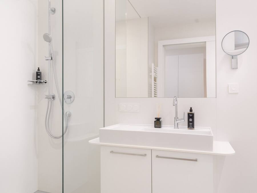 Un baño moderno con cabina de ducha de vidrio, tocador blanco con espejo y un pequeño estante con artículos de aseo.