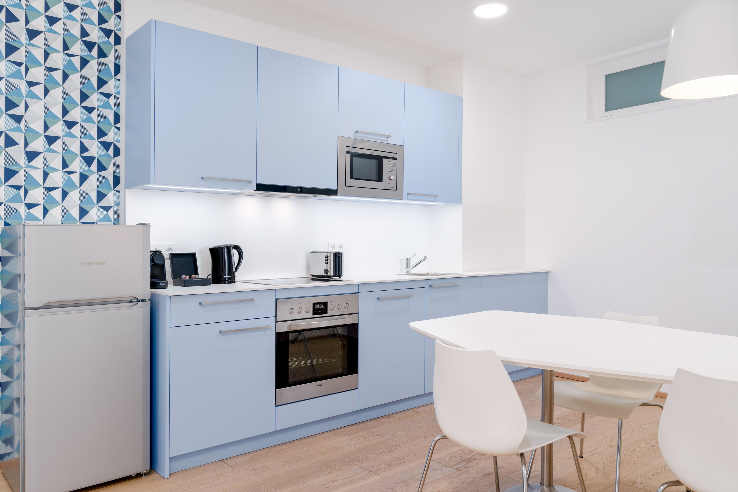 Een moderne keuken met lichtblauwe kastjes, een ingebouwde magnetron, een koffiezetapparaat en een broodrooster. Links staat een witte koelkast en er is een witte eettafel met stoelen.