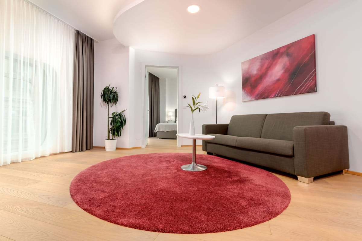 Uma sala de estar moderna com um tapete redondo vermelho, um sofá cinza, uma mesa branca e um quadro abstrato vermelho na parede.