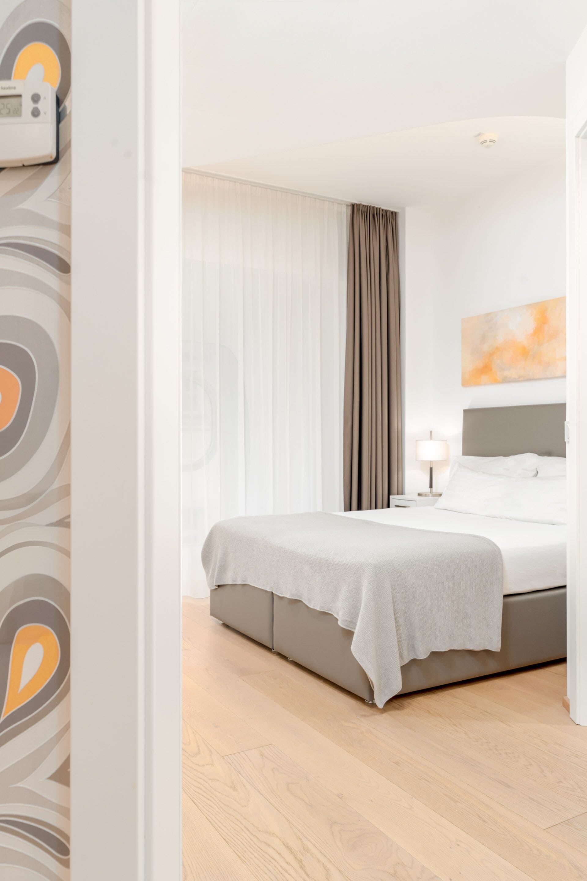 A modern hotel room with a large bed, a bedside table, a lamp, and a view of the outdoors through sheer curtains.