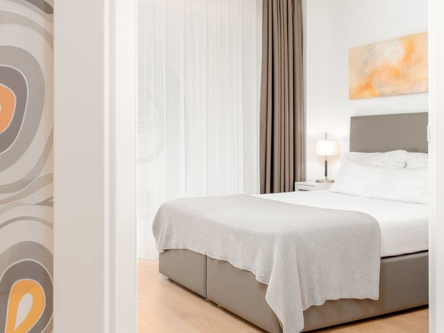 A modern hotel room with a large bed, a bedside table, a lamp, and a view of the outdoors through sheer curtains.