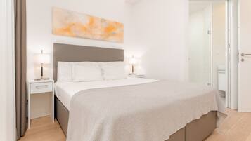 A modern bedroom with a large bed, a bedside table with lamps, a nightstand, and a door leading to another room.