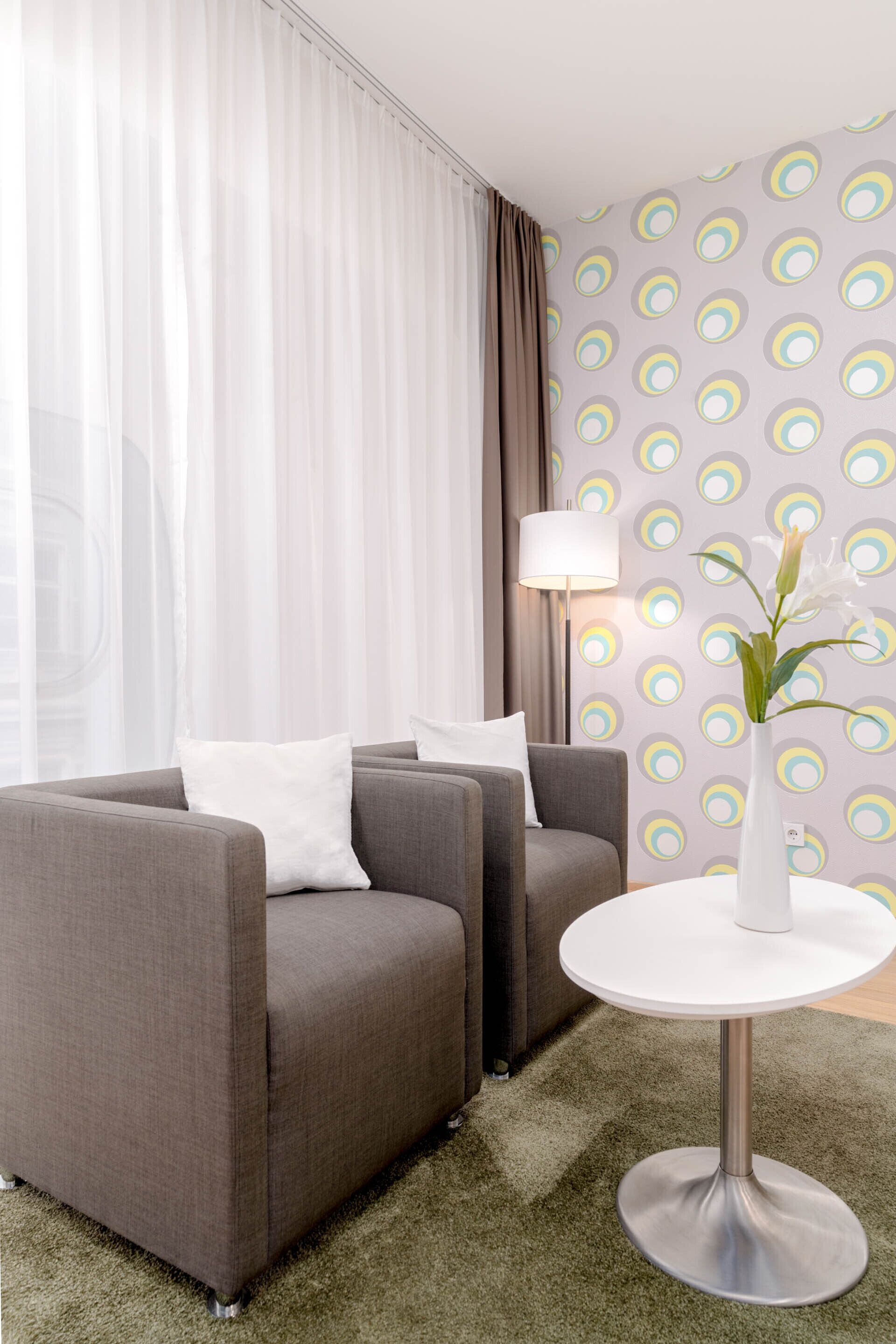 A modern hotel room with a grey sofa set, a round white table, and a patterned wallpaper.