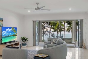 Un soggiorno moderno con una grande TV, una ventola a soffitto e vista su palme e l'oceano.
