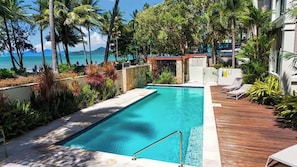 A pool with a wooden deck, surrounded by tropical plants and palm trees, overlooking a body of water.