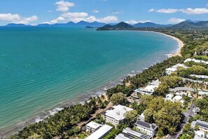 Vista aerea di una zona costiera con spiaggia, edifici e montagne sullo sfondo.