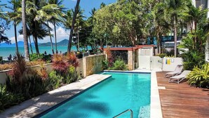 A swimming pool with a wooden deck, surrounded by tropical plants and palm trees, with a view of the ocean and mountains in the background.
