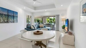 A modern living room with a round dining table, white chairs, a ceiling fan, and a TV on a stand.