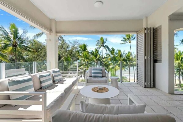 A modern outdoor lounge area with a sofa, chairs, and a coffee table, overlooking a tropical landscape with palm trees.