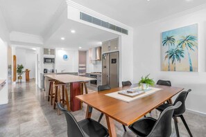 A modern kitchen with a central island, a dining area with a wooden table and black chairs, and a painting of palm trees on the wall.