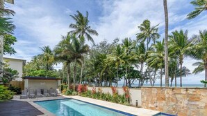 A swimming pool with lounge chairs, surrounded by palm trees and a stone wall, with a view of the ocean. - Turquesa Tides—Beachfront Resort Living (Palm Cove)