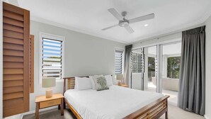 A bedroom with a bed, a ceiling fan, a nightstand, a window with blinds, and a view of the outdoors. - Turquesa Tides—Beachfront Resort Living (Palm Cove)