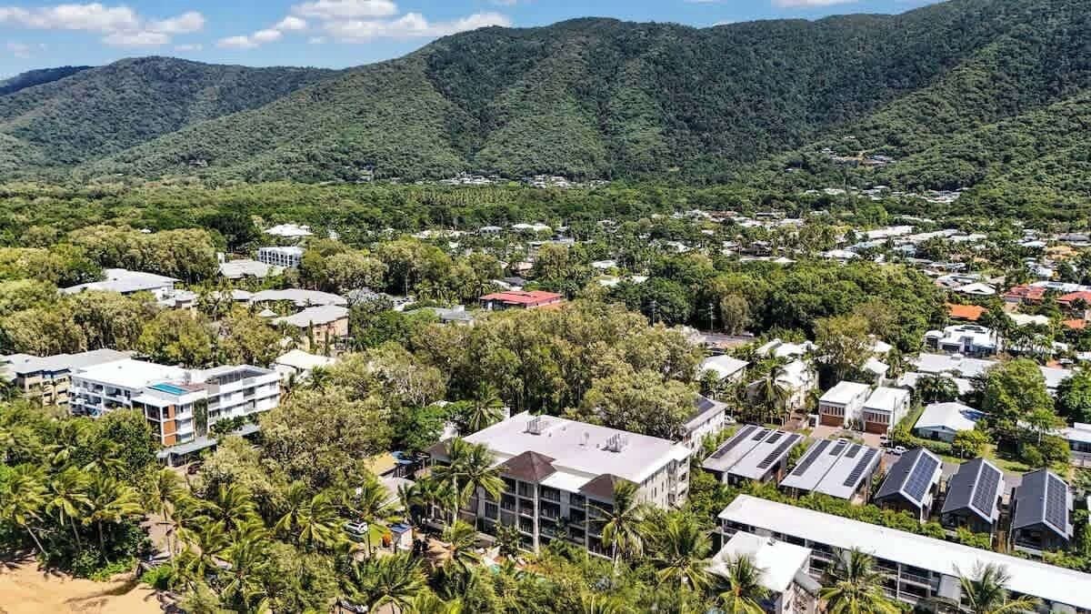 A residential area with houses, some with solar panels, surrounded by dense greenery and hills in the background.