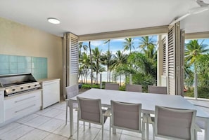 Cucina con piano cottura, tavolo da pranzo e sedie, vista su palme e spiaggia.