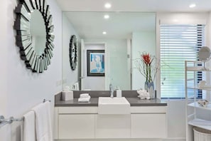 Een moderne badkamer met een grote spiegel, witte kastjes, een wastafel en een vaas met bloemen.