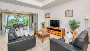 A modern living room with a TV, leather sofas, a wooden coffee table, and a large window with a view of palm trees. - Opal Dreams—Your Gateway to Beachfront Bliss (Palm Cove)