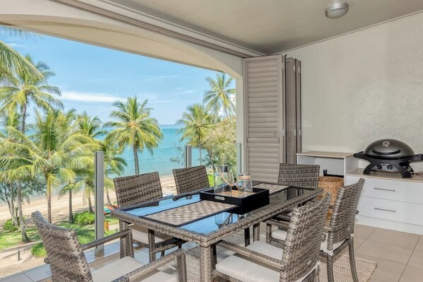 A balcony with a glass table and wicker chairs, overlooking a beach and palm trees. - Opal Dreams—Your Gateway to Beachfront Bliss (Palm Cove)