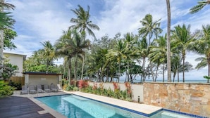 A swimming pool with a stone wall and palm trees, surrounded by a wooden deck and lounge chairs. - Opal Dreams—Your Gateway to Beachfront Bliss (Palm Cove)