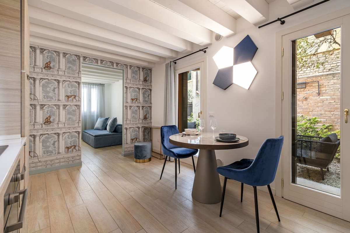 Un espace repas moderne avec une table ronde, des chaises bleues et une vue sur un mur de briques et un jardin, visible à travers une porte vitrée.