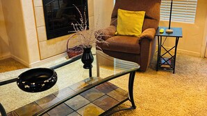 Una sala de estar con chimenea, un sillón marrón con un cojín amarillo, una mesa de centro de vidrio y una lámpara sobre una mesita.