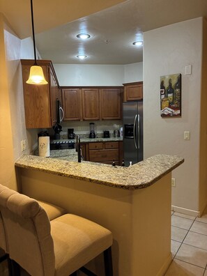 Cocina con armarios de madera, encimeras de granito, refrigerador de acero inoxidable y una zona de bar con una silla.