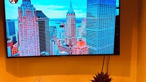 Un televisor montado en la pared muestra una vista urbana. Debajo del televisor, hay un estante con una planta y objetos decorativos.