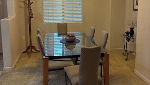 A dining area with a glass table, four chairs, a chandelier, a window with blinds, and a coat rack.