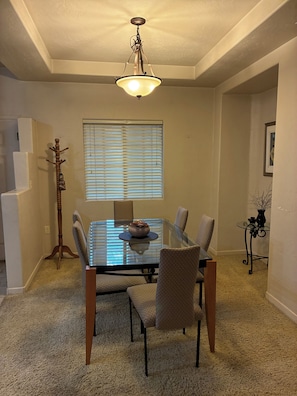 A dining area with a glass table, four chairs, a chandelier, a window with blinds, and a coat rack.