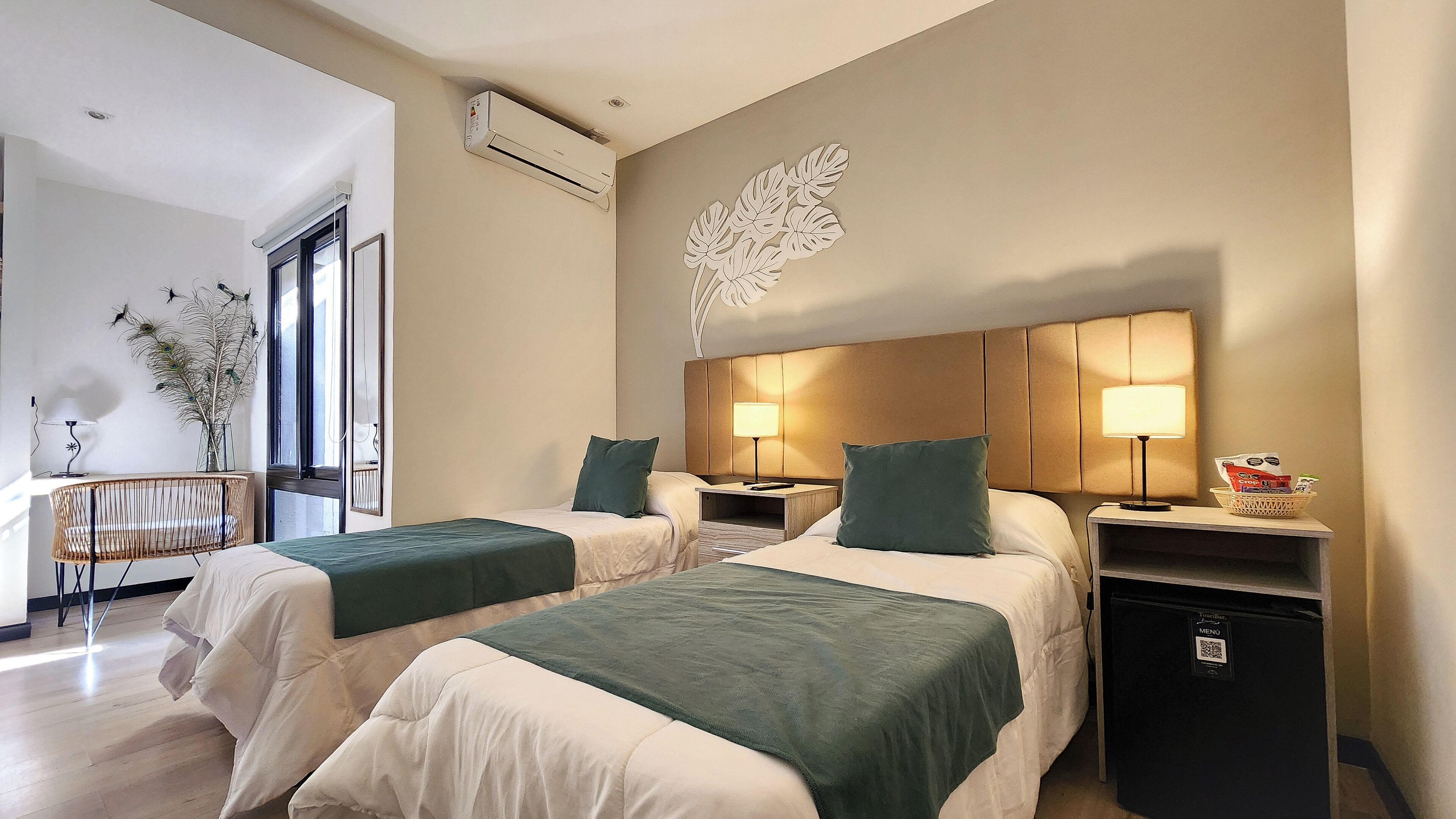 A hotel room with two beds, a wall-mounted air conditioner, a decorative leaf wall art, bedside tables with lamps, and a small basket with items on the nightstand.