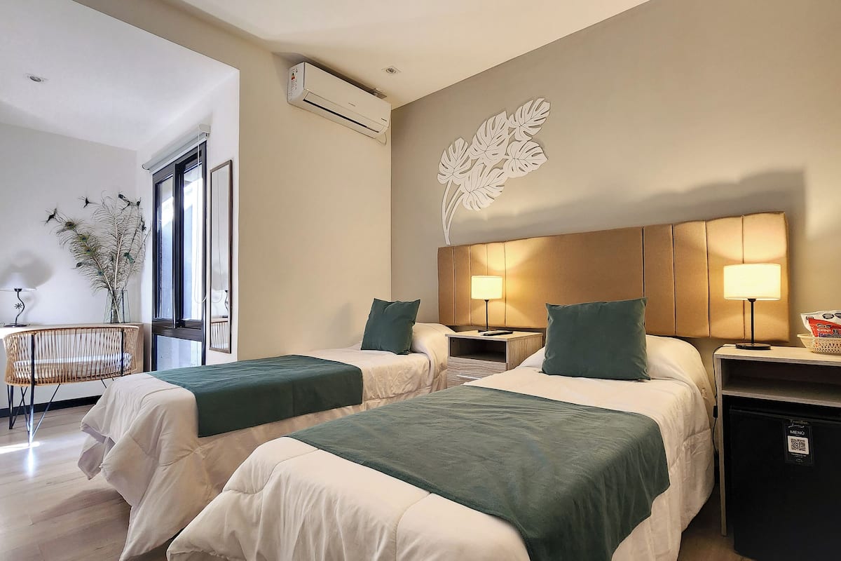 A hotel room with two beds, a wall-mounted air conditioner, a decorative leaf wall art, bedside tables with lamps, and a small basket with items on the nightstand.