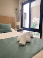 Una cama bien hecha con una colcha verde, un cojín verde azulado y toallas blancas enrolladas.