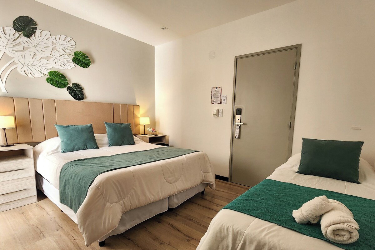 Habitación de hotel con dos camas, suelo de madera, cabecero, mesitas de noche, una lámpara de pared y una puerta.
