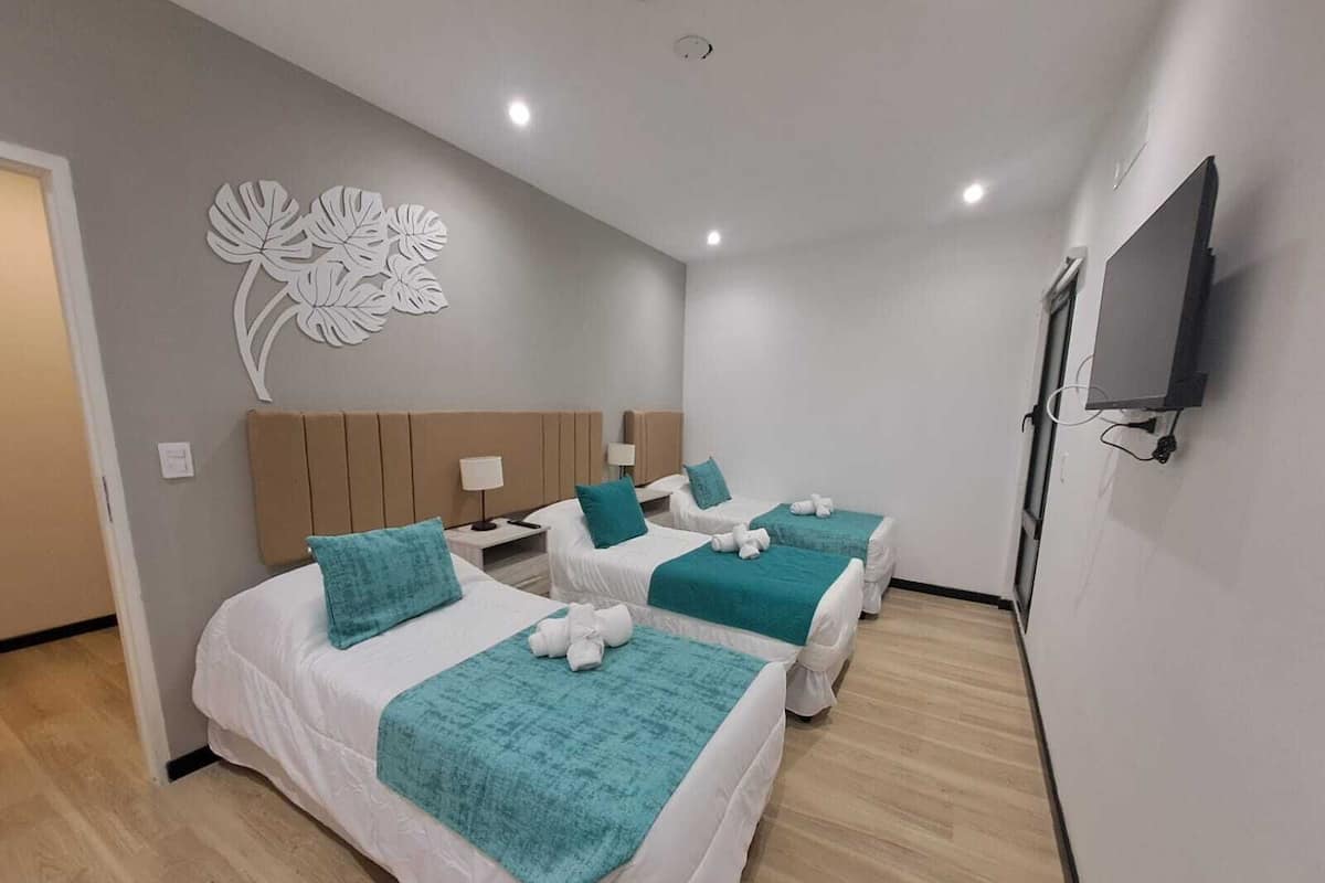 A hotel room with three beds, each with a teal blanket and white pillows. There is a wall-mounted TV and a decorative wall piece.
