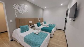 Habitación de hotel con tres camas, cada una con una manta color turquesa y almohadas blancas. Cuenta con un televisor de pared y un adorno mural.