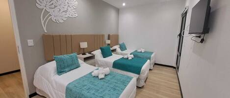 A hotel room with three beds, each with a teal blanket and white pillows. There is a wall-mounted TV and a decorative wall piece.