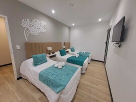 Habitación de hotel con tres camas, cada una con una manta color turquesa y almohadas blancas. Cuenta con un televisor de pared y un adorno mural.