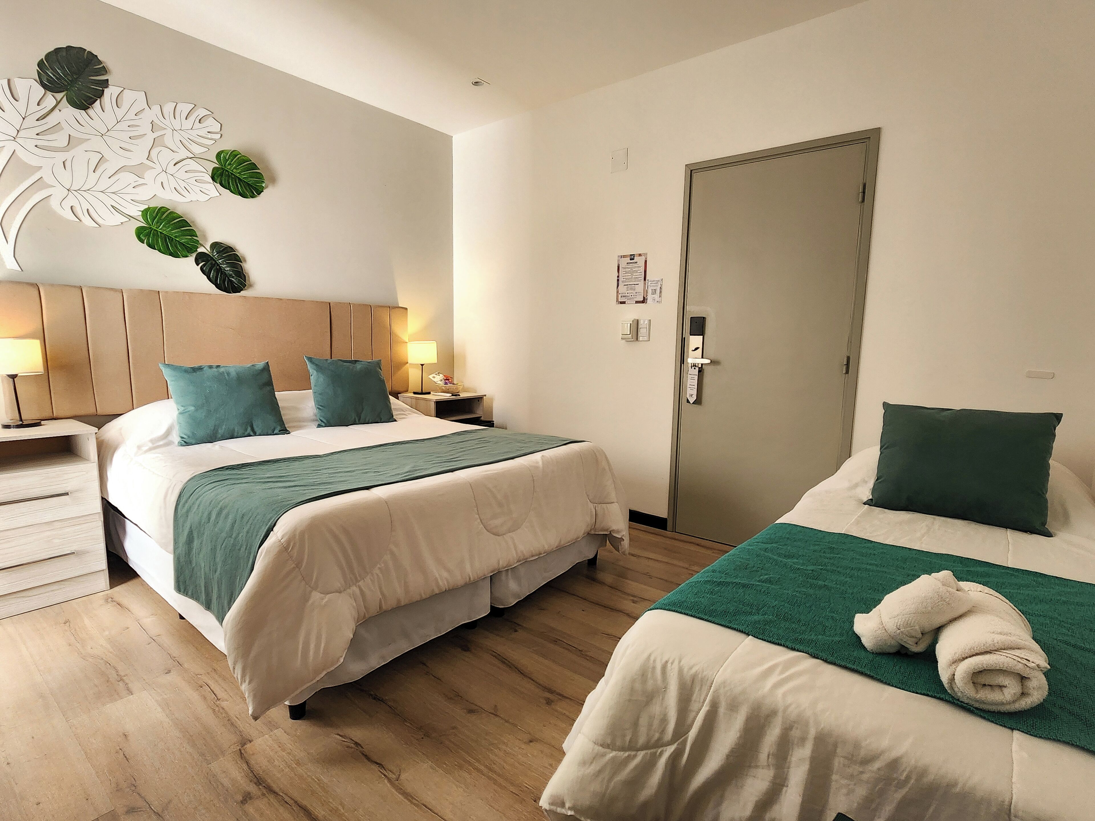 A hotel room with two beds, wooden flooring, a headboard, bedside tables, a wall-mounted light, and a door.