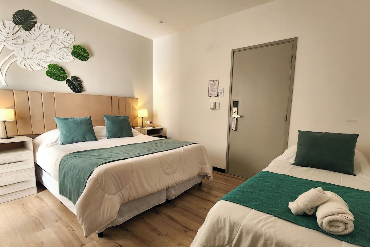 A hotel room with two beds, wooden flooring, a headboard, bedside tables, a wall-mounted light, and a door.