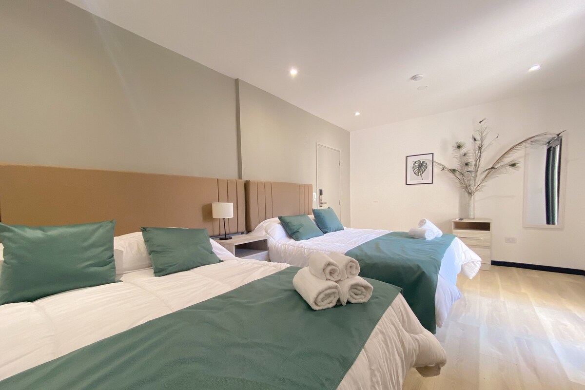 Habitación de hotel con dos camas, cada una con ropa de cama verde y blanca, una mesita de noche con una lámpara y una planta decorativa.