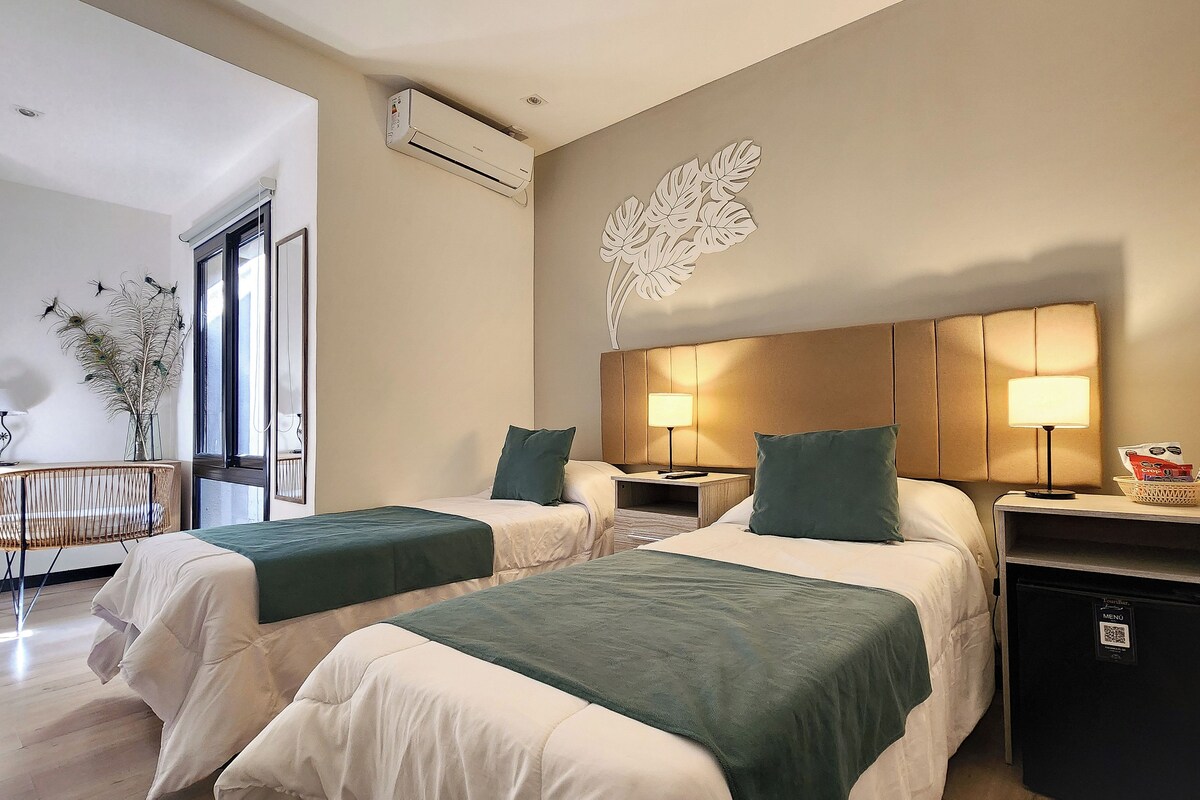 Habitación de hotel con dos camas, aire acondicionado montado en la pared, un cuadro decorativo con hojas, mesitas de noche con lámparas y una pequeña cesta con objetos sobre la mesita.