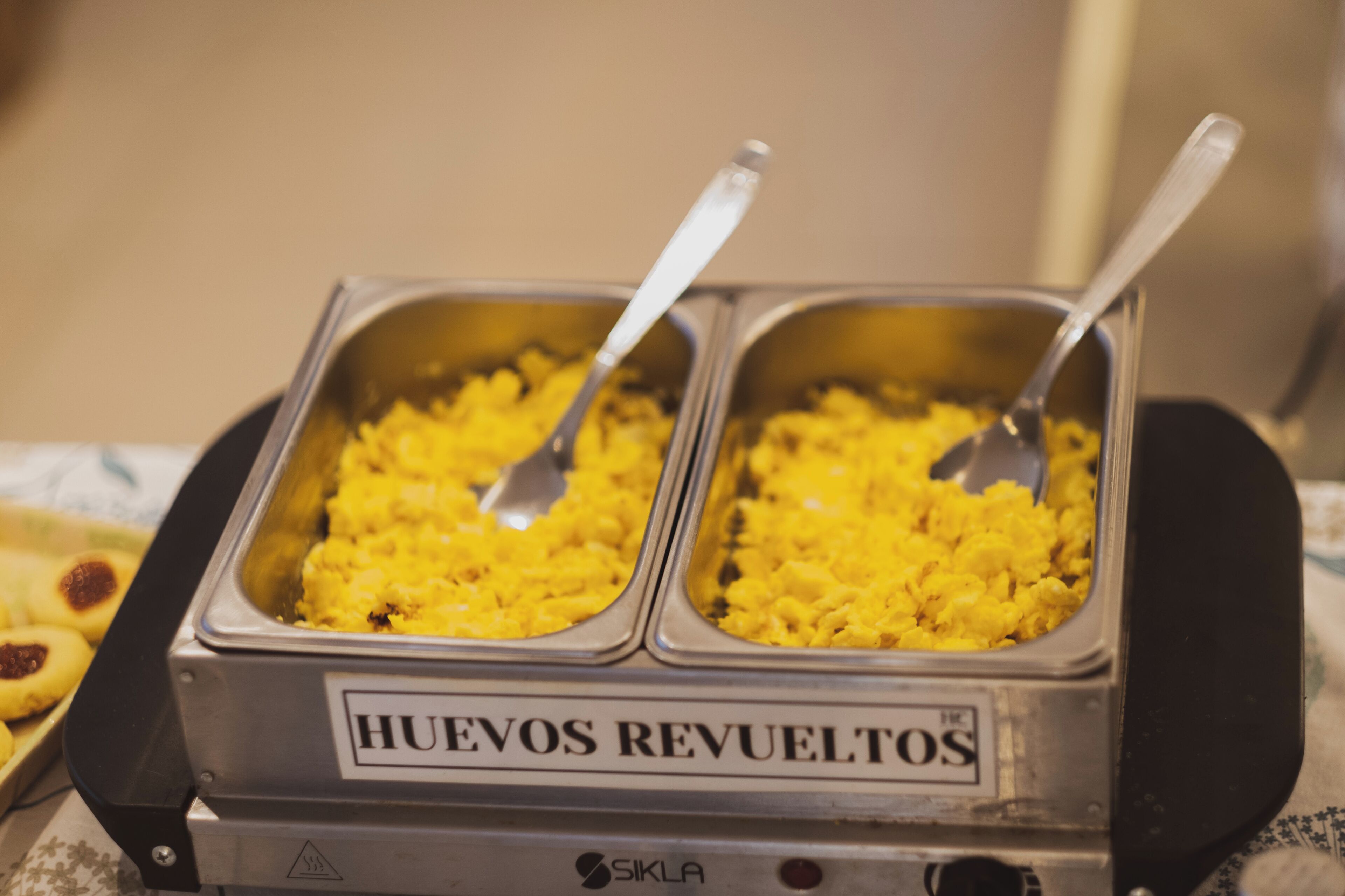 Buffet com dois recipientes rotulados "HUEVOS REVUELTOS" e uma colher em cada um.