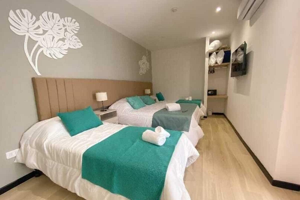 A hotel room with two beds, a wooden headboard, a wall decoration, a television, and a shelf with pillows.