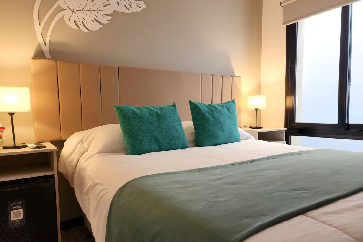 A hotel room with a bed, two green pillows, a white comforter, a green throw blanket, a nightstand with a lamp, and a wall decor piece.