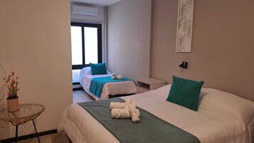 A hotel room with two beds, a small table, a wall-mounted air conditioner, and a window with blinds.