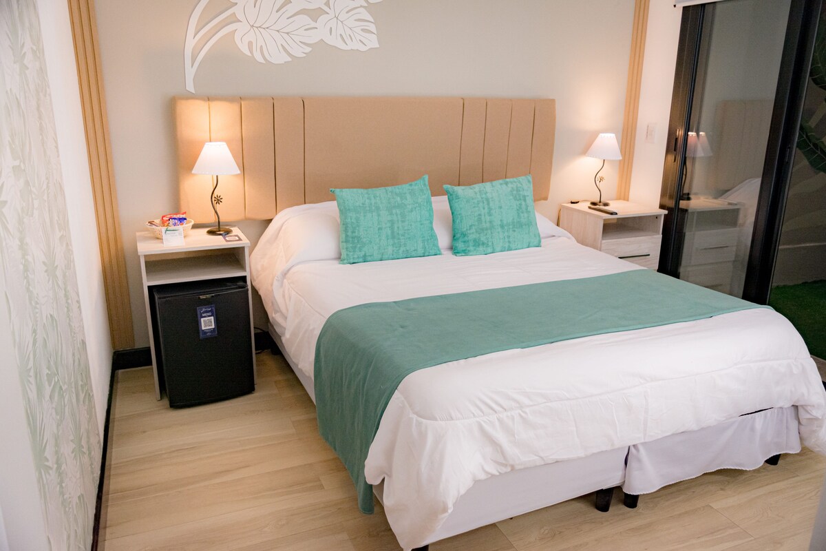 Une chambre d’hôtel comprenant un lit, deux tables de chevet, un petit réfrigérateur et un mur décoré de motifs végétaux.