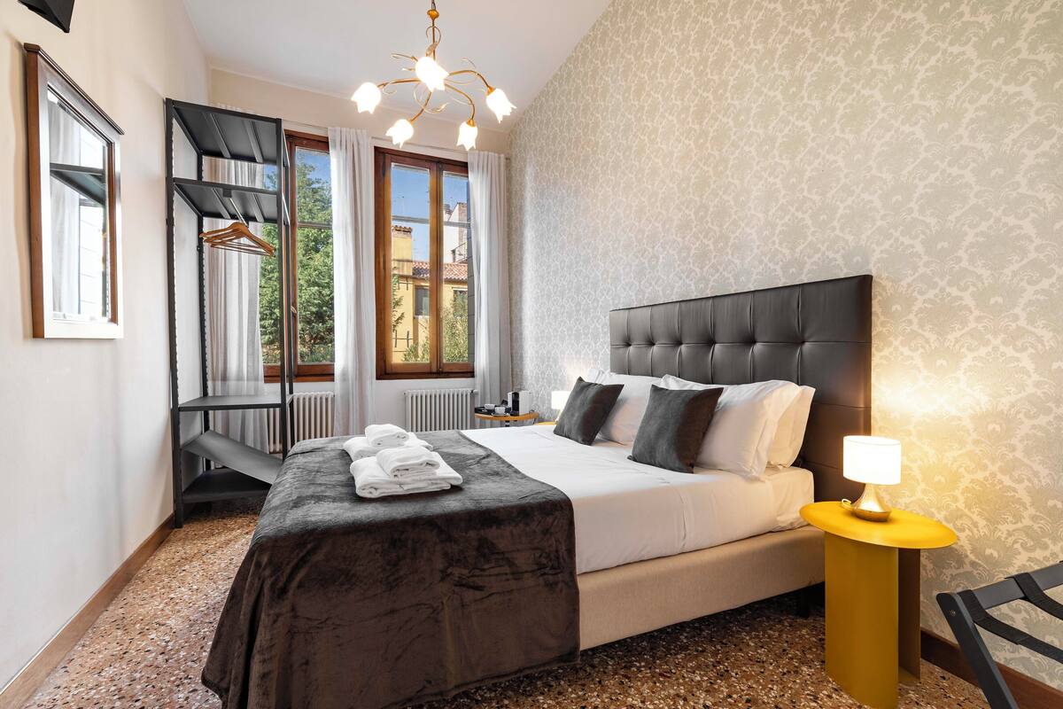 Ein Hotelzimmer mit Bett, Nachttisch mit Lampe, Heizkörper, Fenster mit Vorhängen und einem Kleiderhaken.